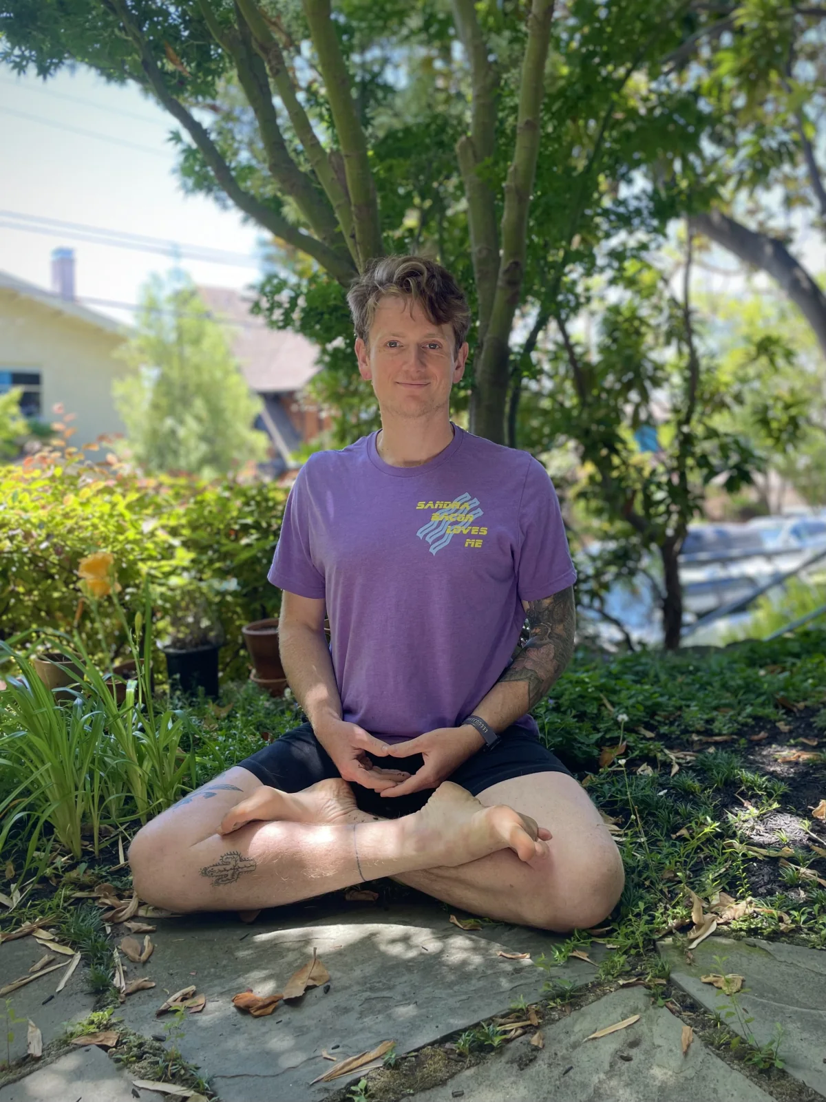Max sitting cross-legged beneath a Japanese maple tree, wearing a purple t-shirt that reads: 'Sandra Bacon Loves Me'.