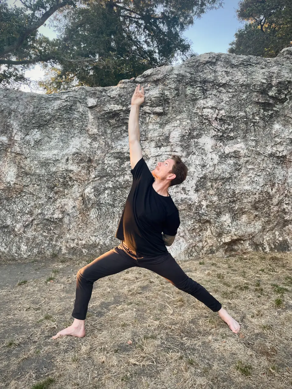 Max in reverse warrior pose, barefoot on dry grass in front of a large rock wall, one arm reaching skyward, wearing black.